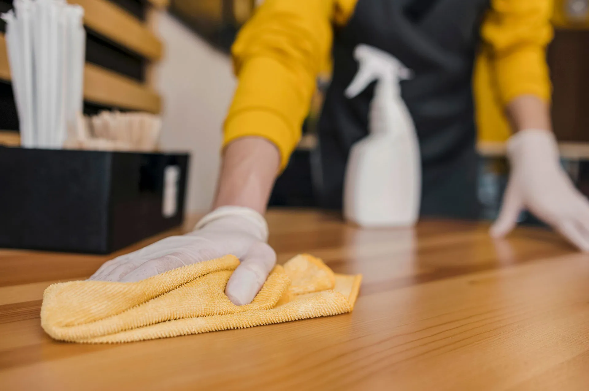 Behandschuhte Hand reinigt eine Holztheke mit Mikrofasertuch und Sprühflasche.